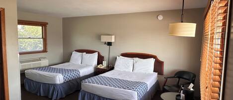 Une chambre d’hôtel avec deux lits, un bureau, une chaise, une lampe et une fenêtre avec des stores.