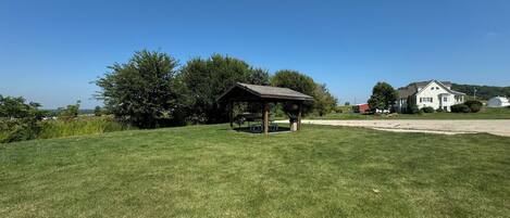 A grassy field with a wooden pavilion, a white house, and a barn in the background.