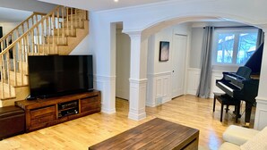 A spacious living room with a wooden staircase, a flat-screen TV on a wooden entertainment unit, a grand piano, and a large window with a view of trees.