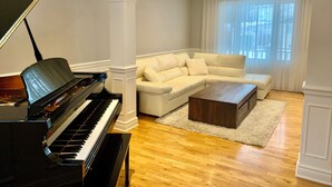 A living room with a grand piano, a white sectional sofa, a wooden coffee table, and a large window with sheer curtains.