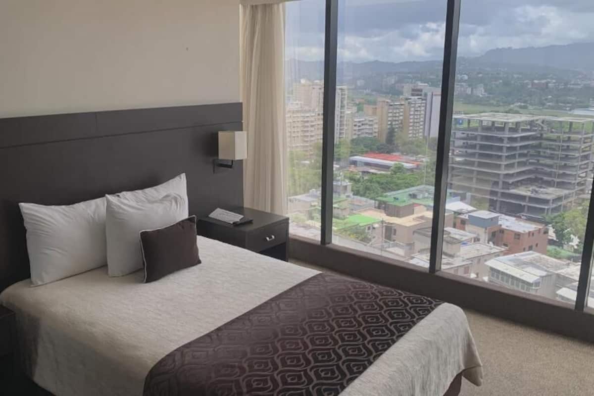 Habitación de hotel con una cama grande, una mesita de noche, una lámpara y vistas a un paisaje urbano a través de amplios ventanales.