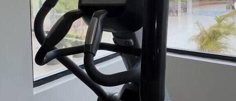 Una bicicleta estática con pantalla digital y manillares, ubicada cerca de una ventana con vista a un edificio y follaje.