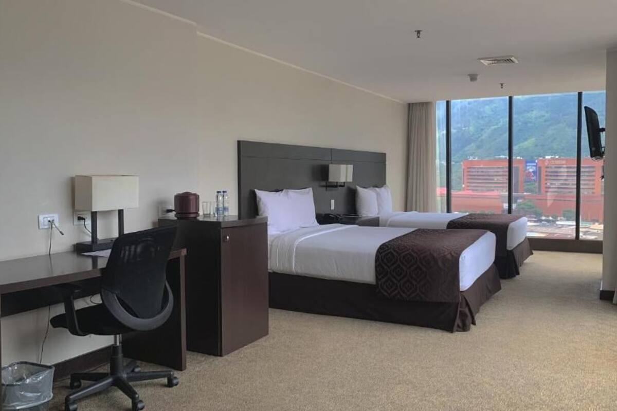 Habitación de hotel con dos camas, un escritorio con silla, televisor y un amplio ventanal con vistas a edificios.