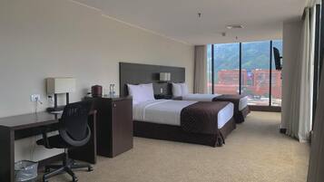 A hotel room with two beds, a desk with a chair, a TV, and a large window with a view of buildings.
