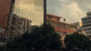 Um arranha-céu alto e moderno com fachada de vidro refletante, rodeado por outros edifícios e árvores.