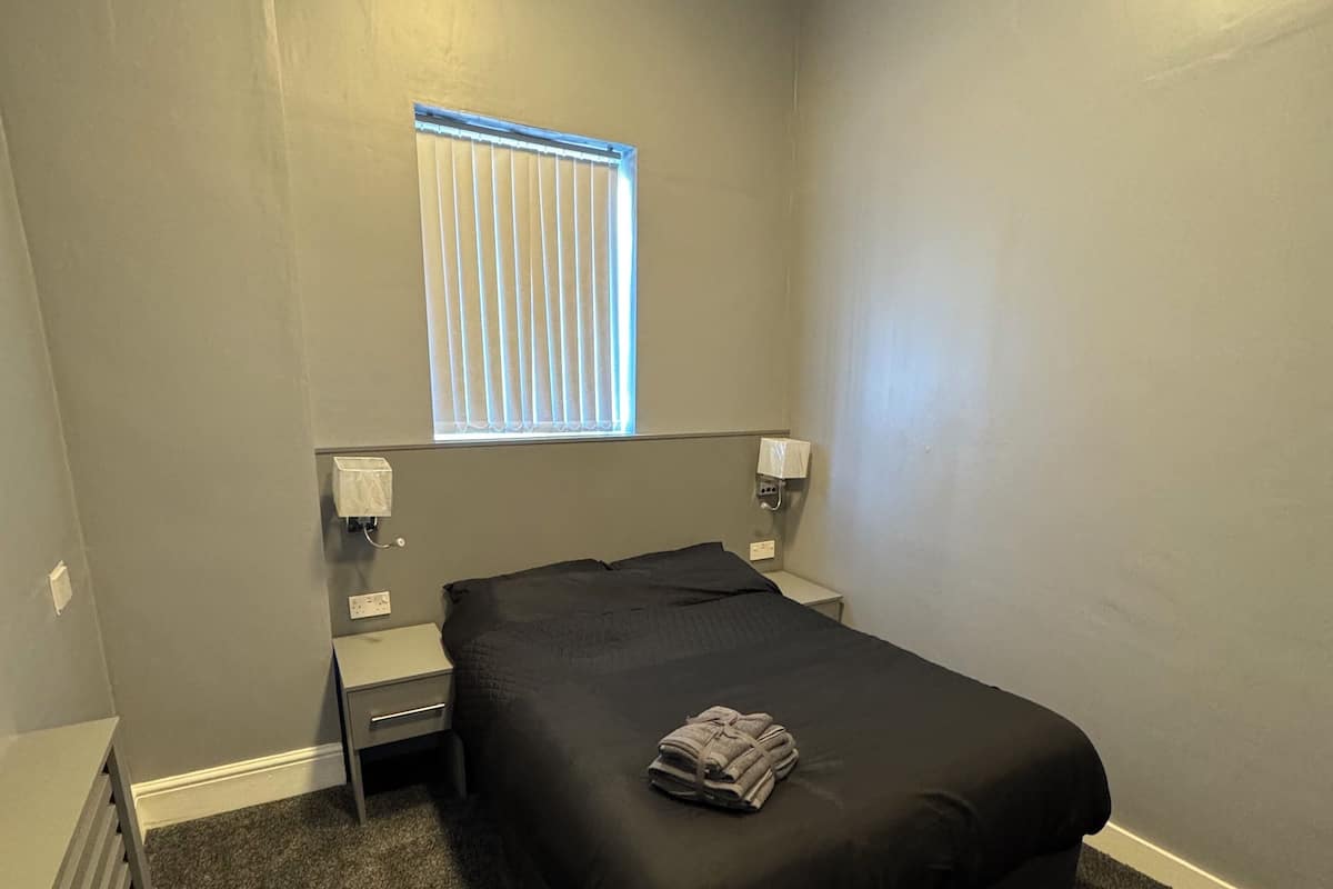 A bedroom with a single bed, a nightstand, a wall socket, and a window with blinds.