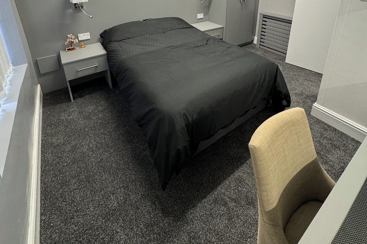 A hotel room with a bed, bedside table, lamp, wardrobe, and a chair.