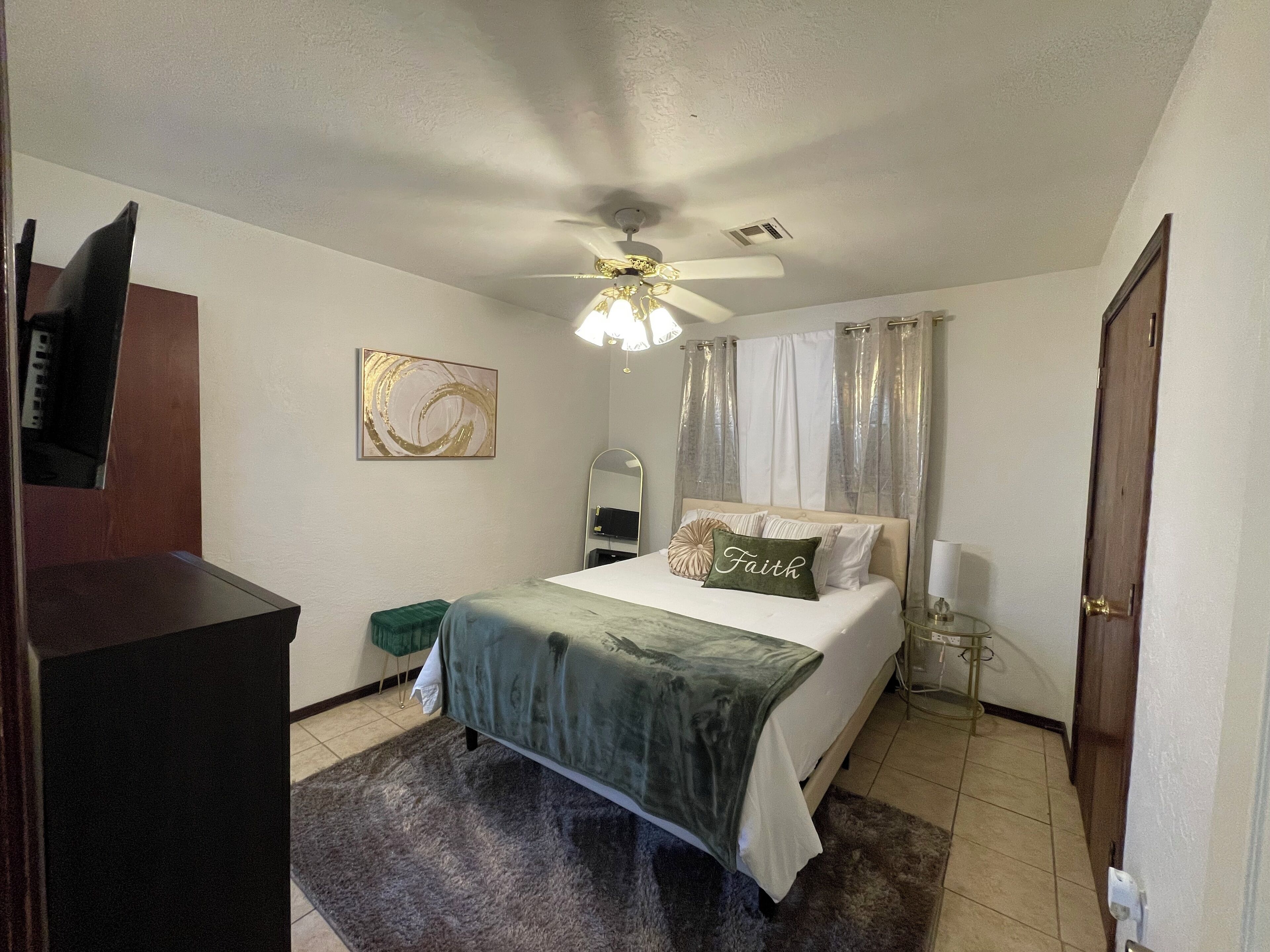 Una camera d'albergo con un letto, una ventola a soffitto, un comodino, uno specchio, una sedia verde e una TV fissata al muro.
