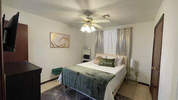 Una camera d'albergo con un letto, una ventola a soffitto, un comodino, uno specchio, una sedia verde e una TV fissata al muro.