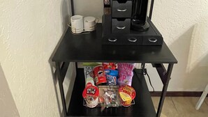 A black metal shelf unit with multiple compartments, including a top shelf with decorative jars, a middle shelf with a glass container and office supplies, and a bottom shelf with snack packages. - Lawton/Fort Sill Home near Airport  (Lawton)