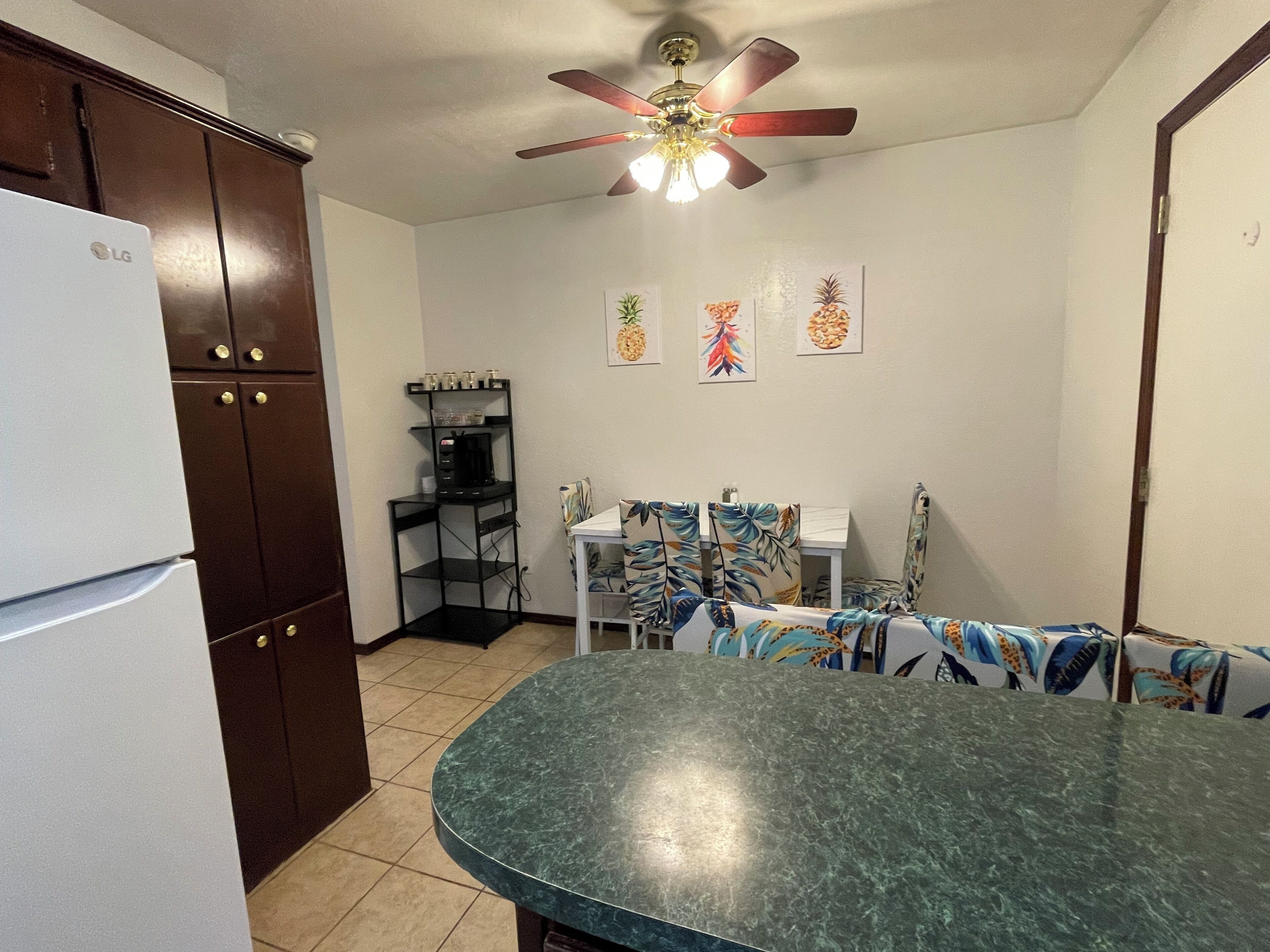 Una cocina con refrigerador, ventilador de techo, cafetera en estante y una mesa de comedor con sillas.