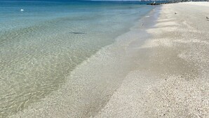 A beach with clear shallow waters, a sandy shore, and a distant shoreline with buildings.
