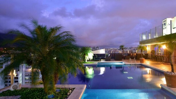 A pool area with a view of a cityscape at dusk, palm trees, and a modern building with a lounge area. - Hotel Pestana Caracas Premium (Caracas)