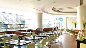 A restaurant with a modern interior, featuring a mix of booth and table seating, large windows offering a view of the city, and a variety of plants and flowers as decor. - Hotel Pestana Caracas Premium (Caracas)