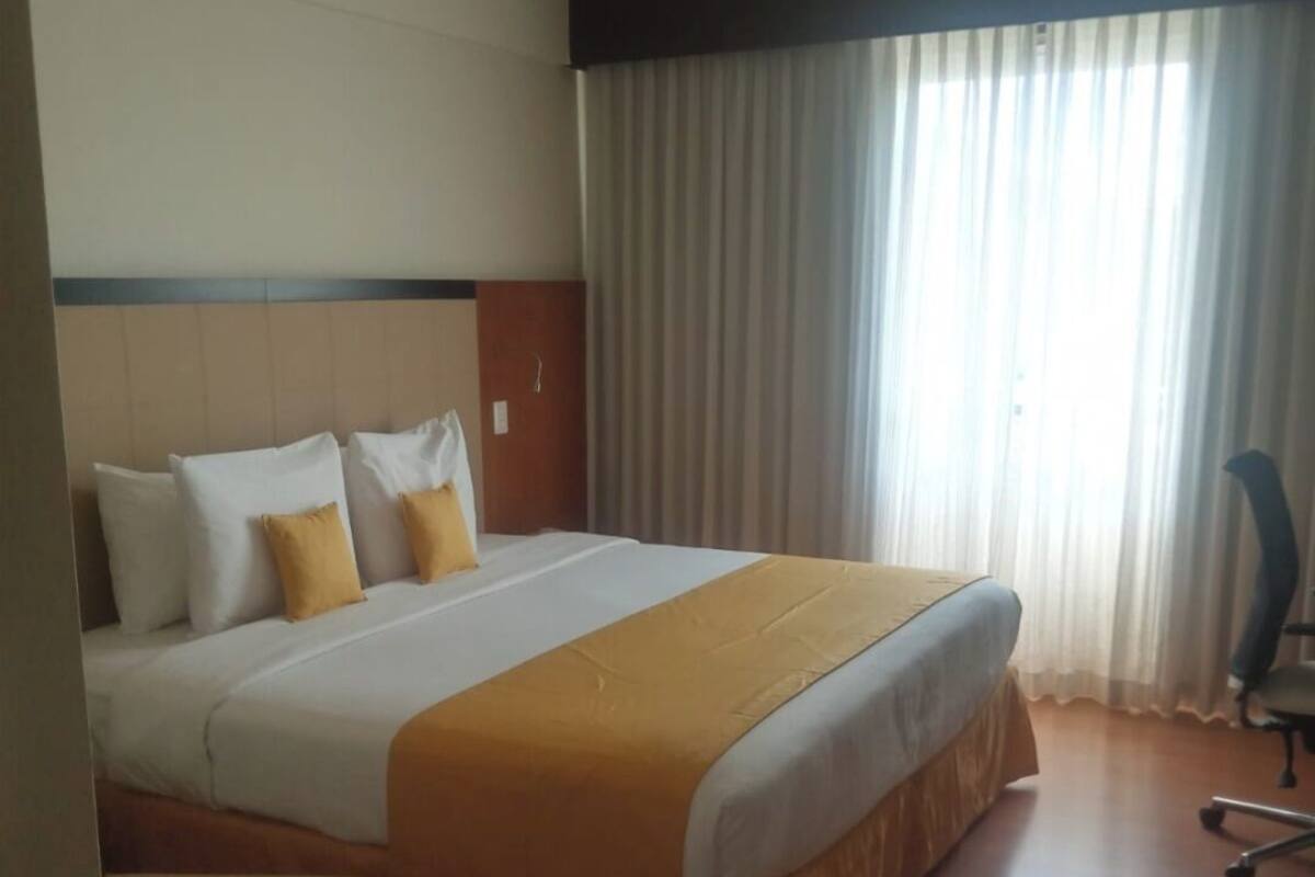 Ein Hotelzimmer mit einem großen Bett, einem Holzkopfende, einem Fenster mit Vorhängen und einem Sessel.