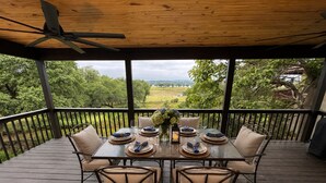 Un área de comedor al aire libre techada con una mesa dispuesta para cuatro personas, rodeada de sillas, y vistas a la vegetación y colinas al fondo.