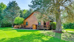 A rustic log cabin with a wooden deck, surrounded by lush greenery and trees.