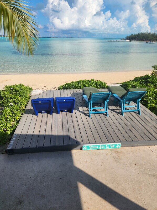 Un pontile in legno con poltrone da relax blu e un tavolo, con vista su una spiaggia e un mare turchese.