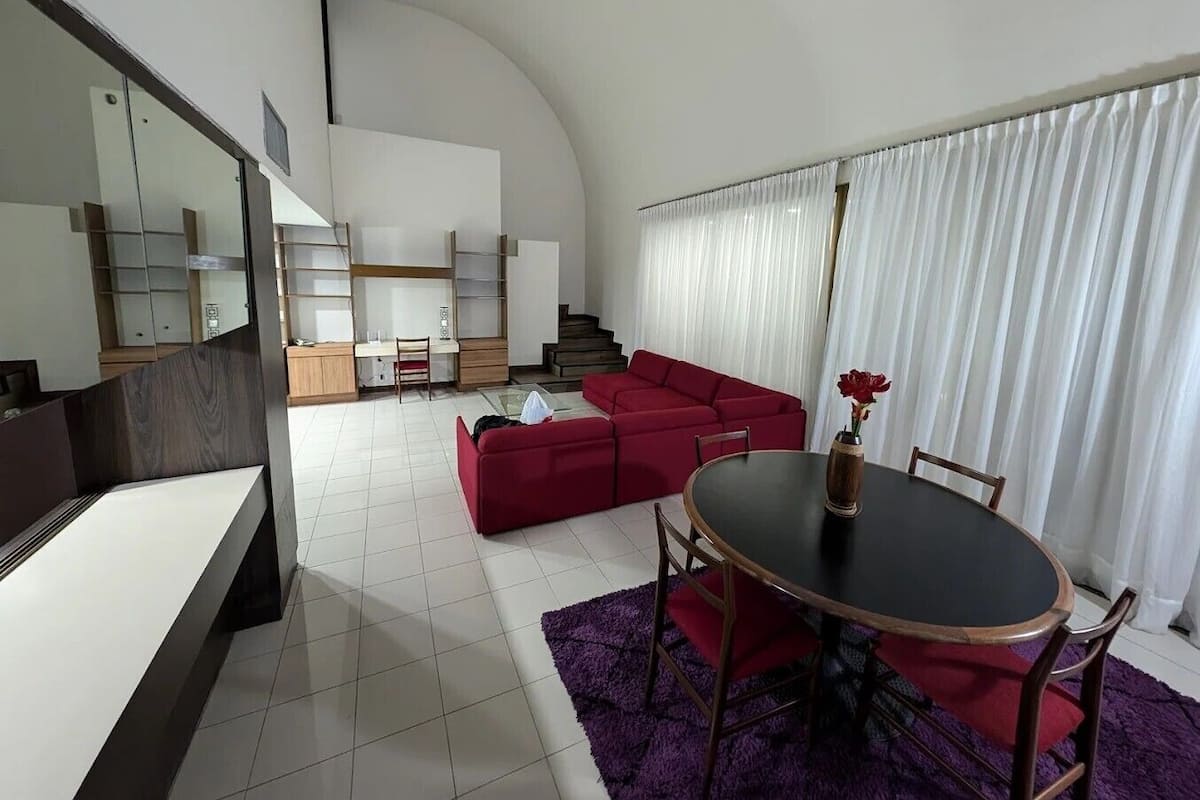 Una sala de estar moderna con un sofá rojo, una mesa de comedor con sillas y una escalera que sube a una zona de entrepiso.