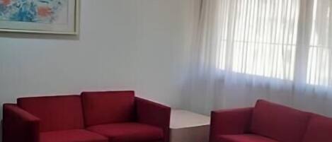 A living room with two red sofas and a white coffee table. There is a painting on the wall and a window with sheer curtains.