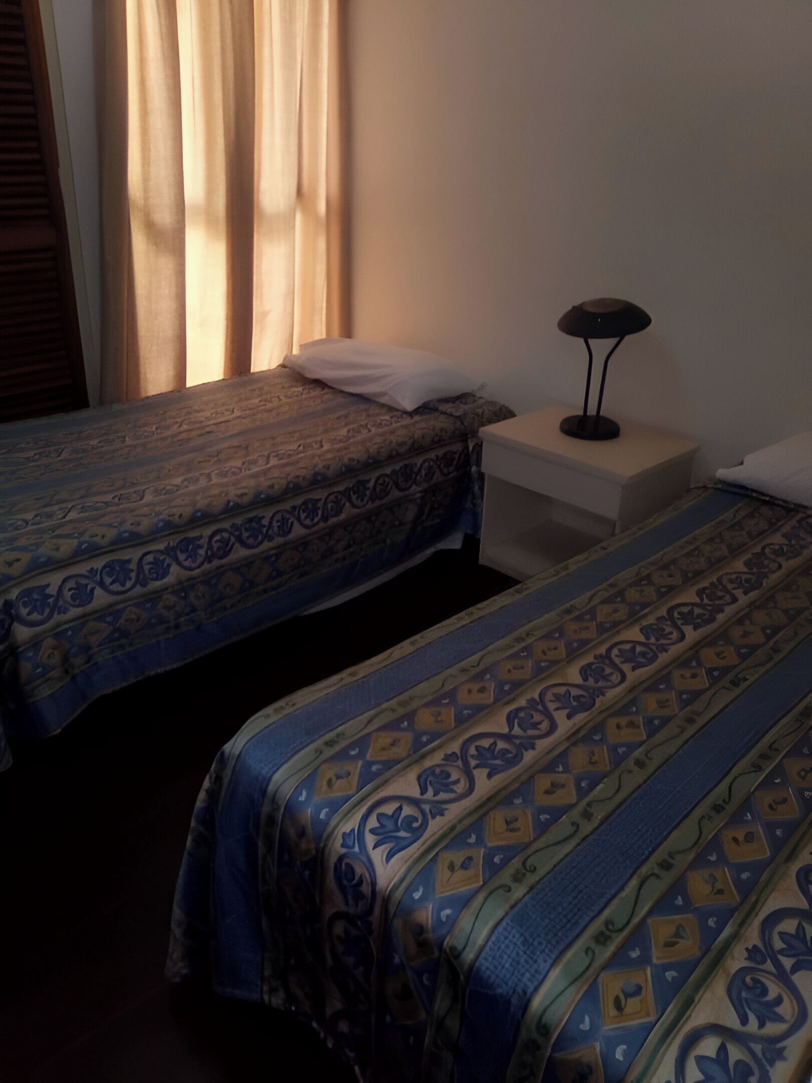 A room with two beds, each with patterned bedding, a bedside table with a lamp, and a window with curtains.