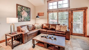 A spacious living room with a leather sectional sofa, a coffee table with candles, a floor lamp, and a large window with blinds. - Ski-in 2 Bedroom Townhome at Mountain Thunder (Breckenridge)