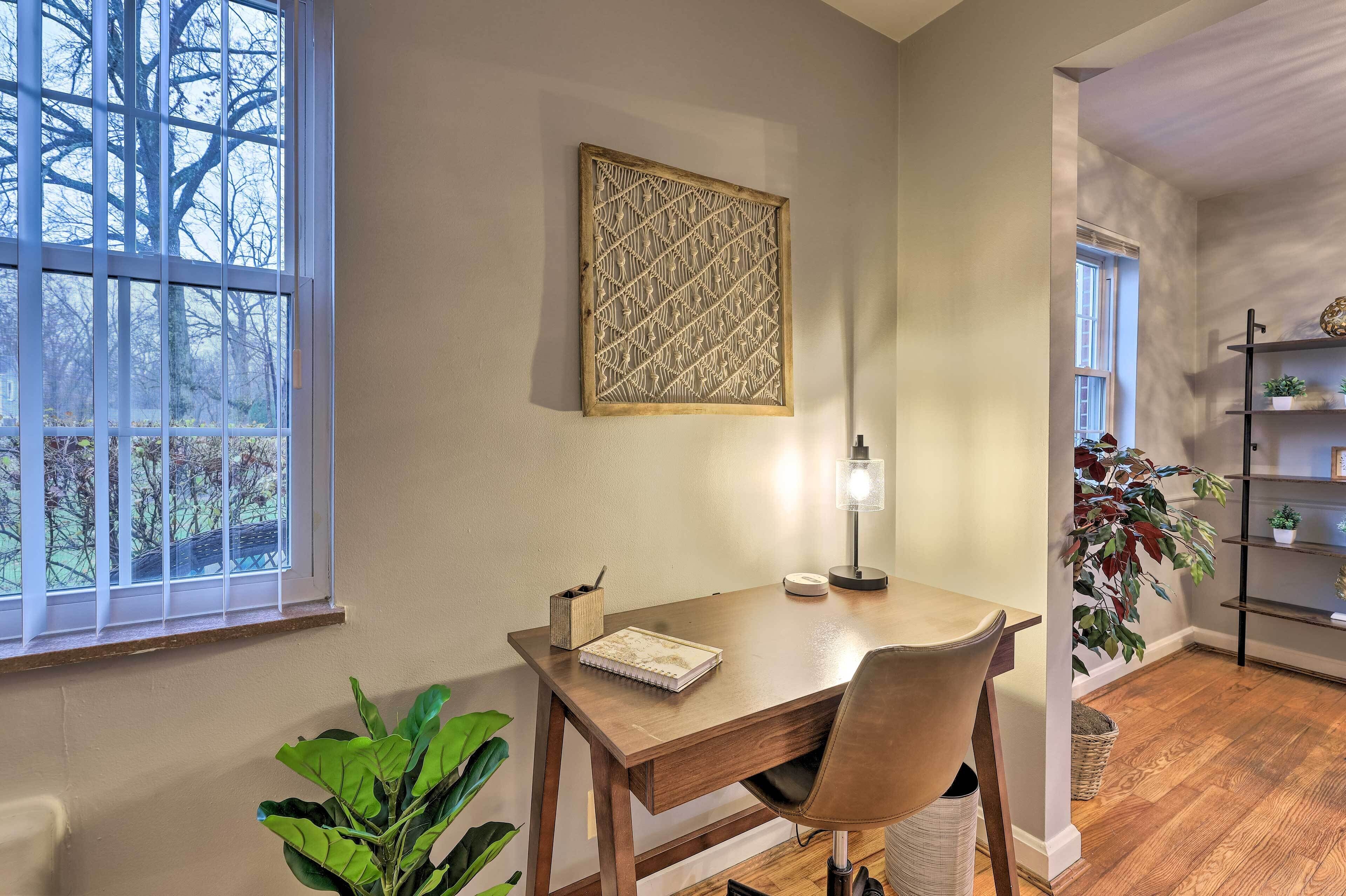 A room with a wooden desk, a chair, a lamp, a framed artwork, and a bookshelf with plants.