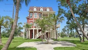 Una casa grande de dos pisos con fachada roja y detalles blancos, rodeada de abundante follaje y palmeras.