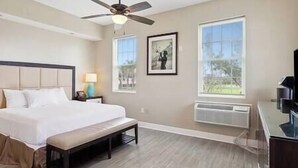 A hotel room with a bed, a ceiling fan, two windows, a framed picture, and a desk with a lamp. - Magnolia Cottage R2 (Lafitte)