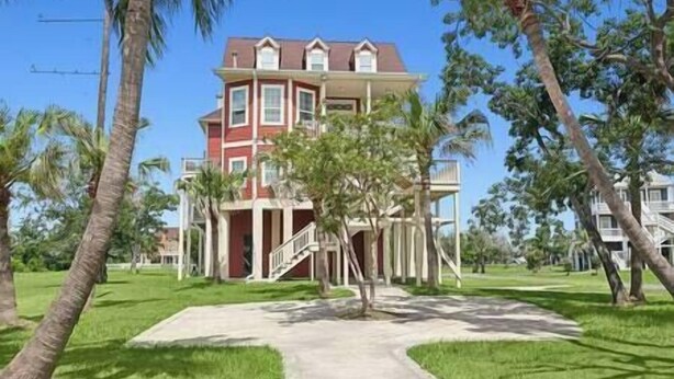 A two-story building with a red facade, a central entrance with a staircase, and a surrounding lawn with palm trees. - Oak Cottage R103 (Lafitte)