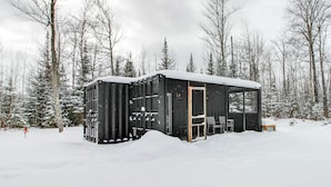 Une cabane noire, située dans une forêt enneigée, avec une porte et un banc à l’extérieur.