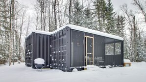 En modern, svart stuga med ett stort fönster och en dörr, omgiven av snötäckta träd.