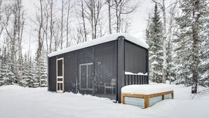 Una cabaña nevada con un amplio ventanal corredera, un banco exterior y vista a un bosque cubierto de nieve.