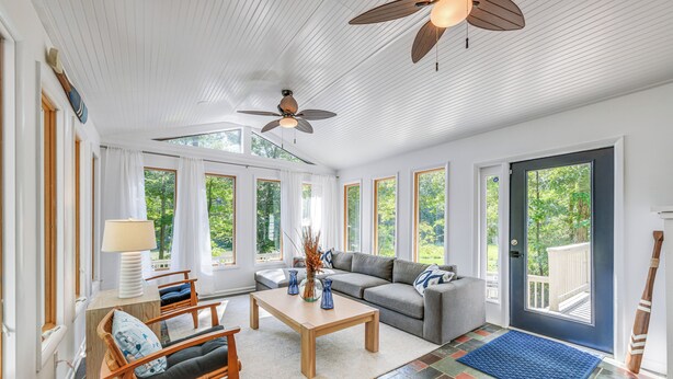 A spacious living room with a sofa, coffee table, and chairs, large windows, and a ceiling fan. - Waterfront Urbanna Home w/ Private Dock (Urbanna)