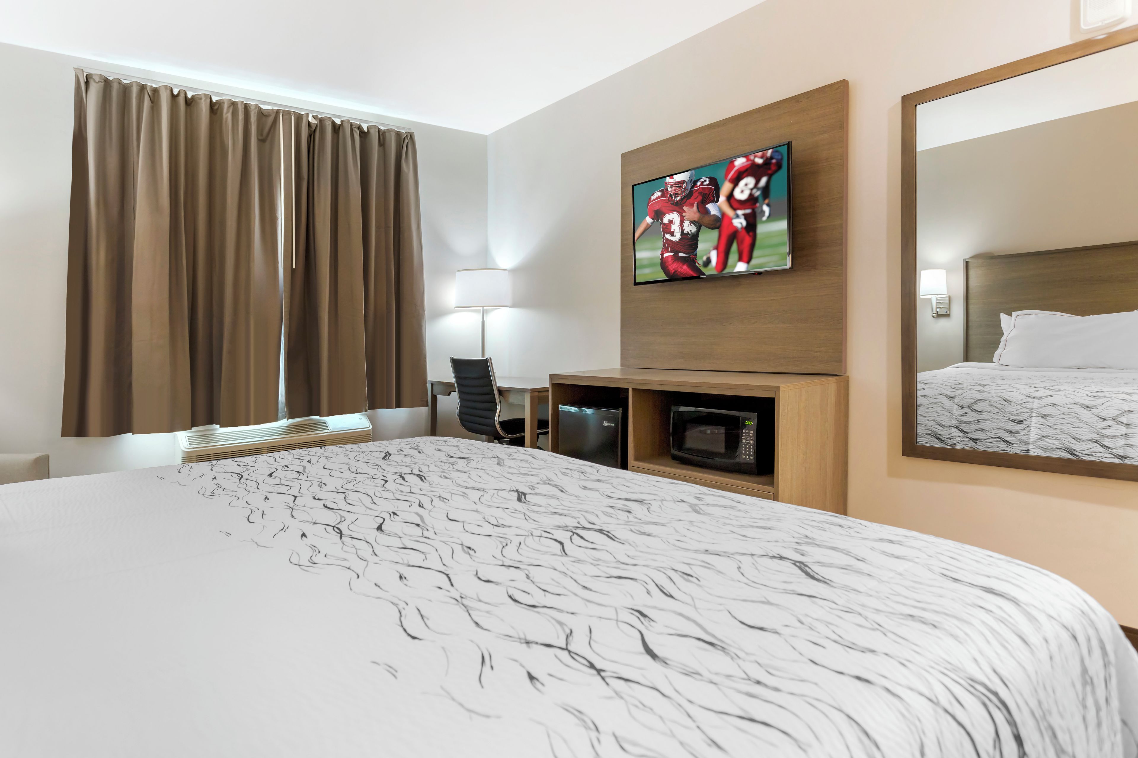 A hotel room with a large bed, a flat-screen TV mounted on the wall, a desk with a chair, a microwave, and a large mirror.