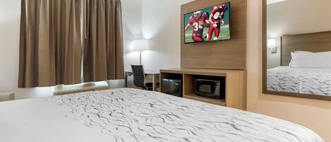 A hotel room with a large bed, a flat-screen TV mounted on the wall, a desk with a chair, a microwave, and a large mirror.