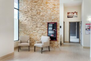 Un pasillo con pared de piedra, dos sillas, un panel de control de alarma contra incendios y una puerta que da a otra habitación.