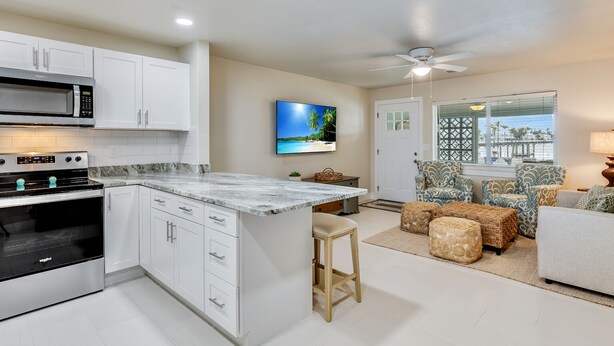 A modern kitchen with white cabinets, a built-in oven, and a microwave. There is a dining area with patterned chairs and a view of the beach through the window. - Turtle Cove at Siesta Dreams - private 2BR, 1BA apartment near Fort Myers Beach! (Fort Myers Beach)
