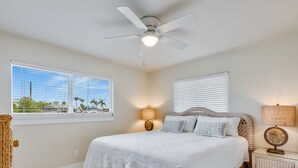 A bedroom with a large bed, a ceiling fan, two bedside lamps, a wicker nightstand, and a view of palm trees through the window. - Turtle Cove at Siesta Dreams - private 2BR, 1BA apartment near Fort Myers Beach! (Fort Myers Beach)