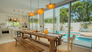 Et moderne køkken med en stor ø, et træspisebord og bænk, med udsigt til en swimmingpool og grønne omgivelser.