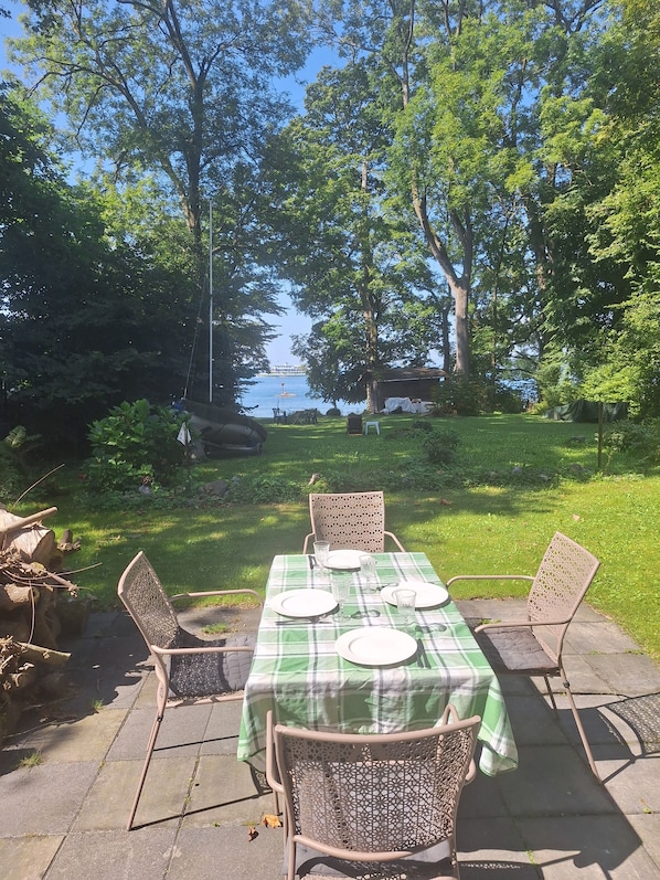 An outdoor dining area with a table set for four, surrounded by chairs, set against a backdrop of trees and a view of the water.