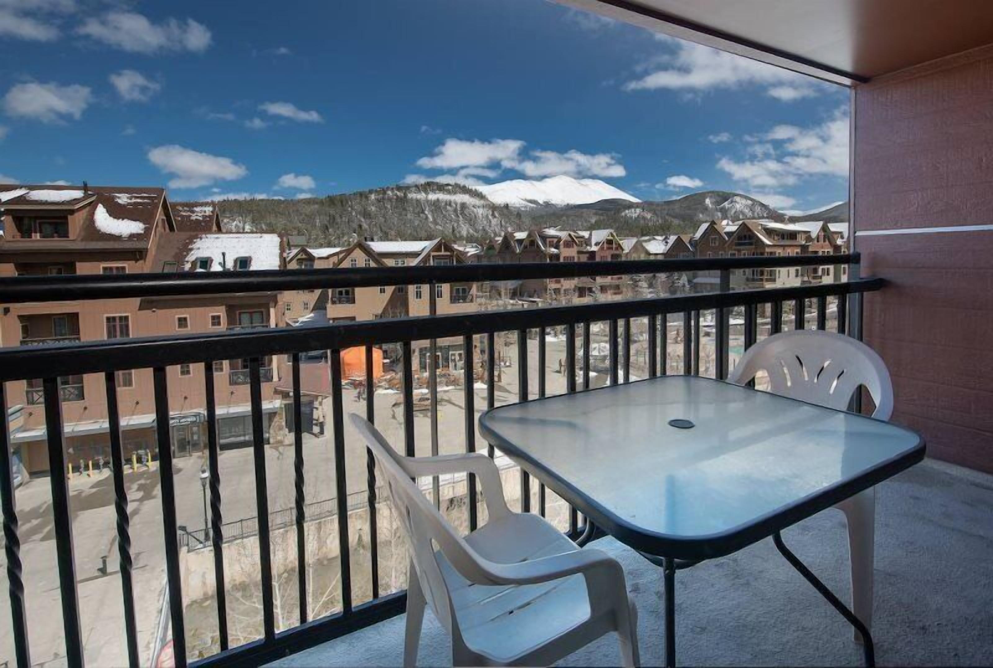 A balcony with a table and two chairs, overlooking a residential area with snow-capped mountains in the background.