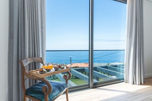 A modern room with a large window, a wooden chair with a tray of breakfast, and a view of the sea and buildings. - Sapphire Sands by Atlantic Holiday (Calheta)