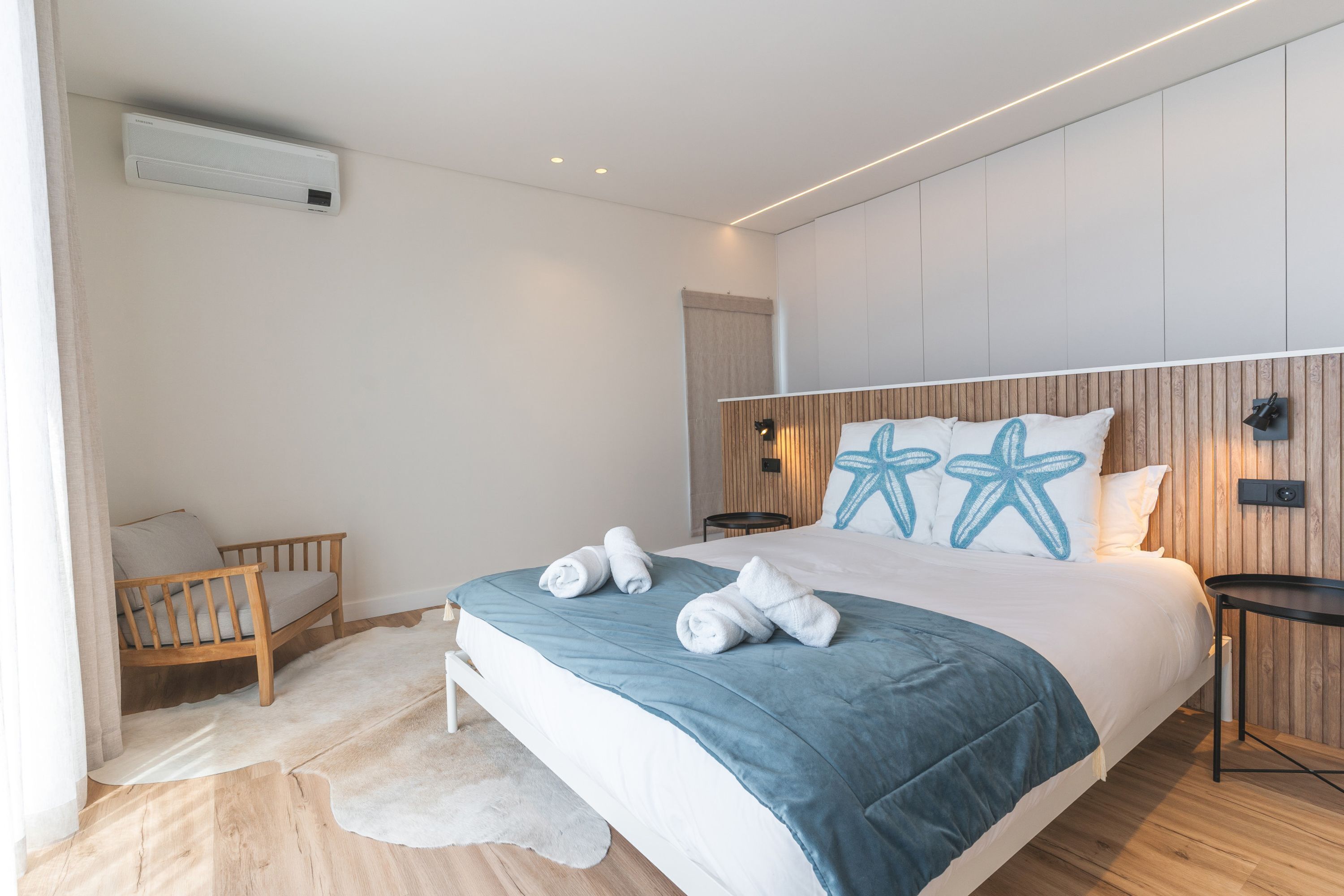 A modern bedroom with a bed, a wooden bench, a small table, and a wall-mounted air conditioner.