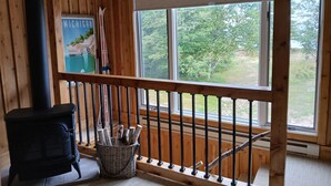 A wooden cabin interior with a black wood stove, a window with a view of greenery, and a poster on the wall. - Rustic Moose Lakefront Guest Cabin on the Shore of Lake Superior (Brimley)