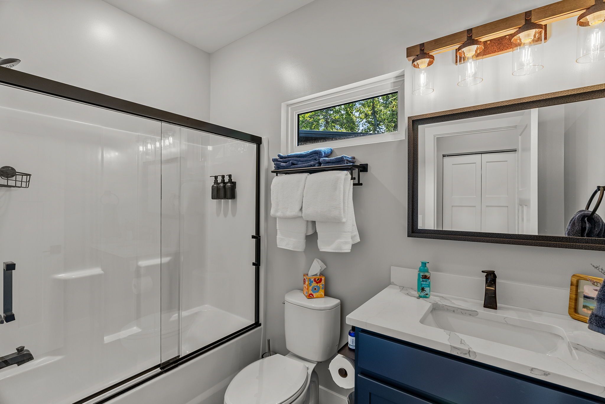 A modern bathroom with a glass-enclosed shower, a toilet, a sink with a mirror above, and a window providing natural light.