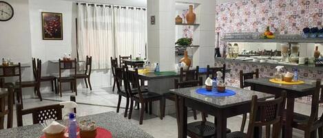 A dining area with multiple tables and chairs, a kitchen area with tiled backsplash, and a clock on the wall.