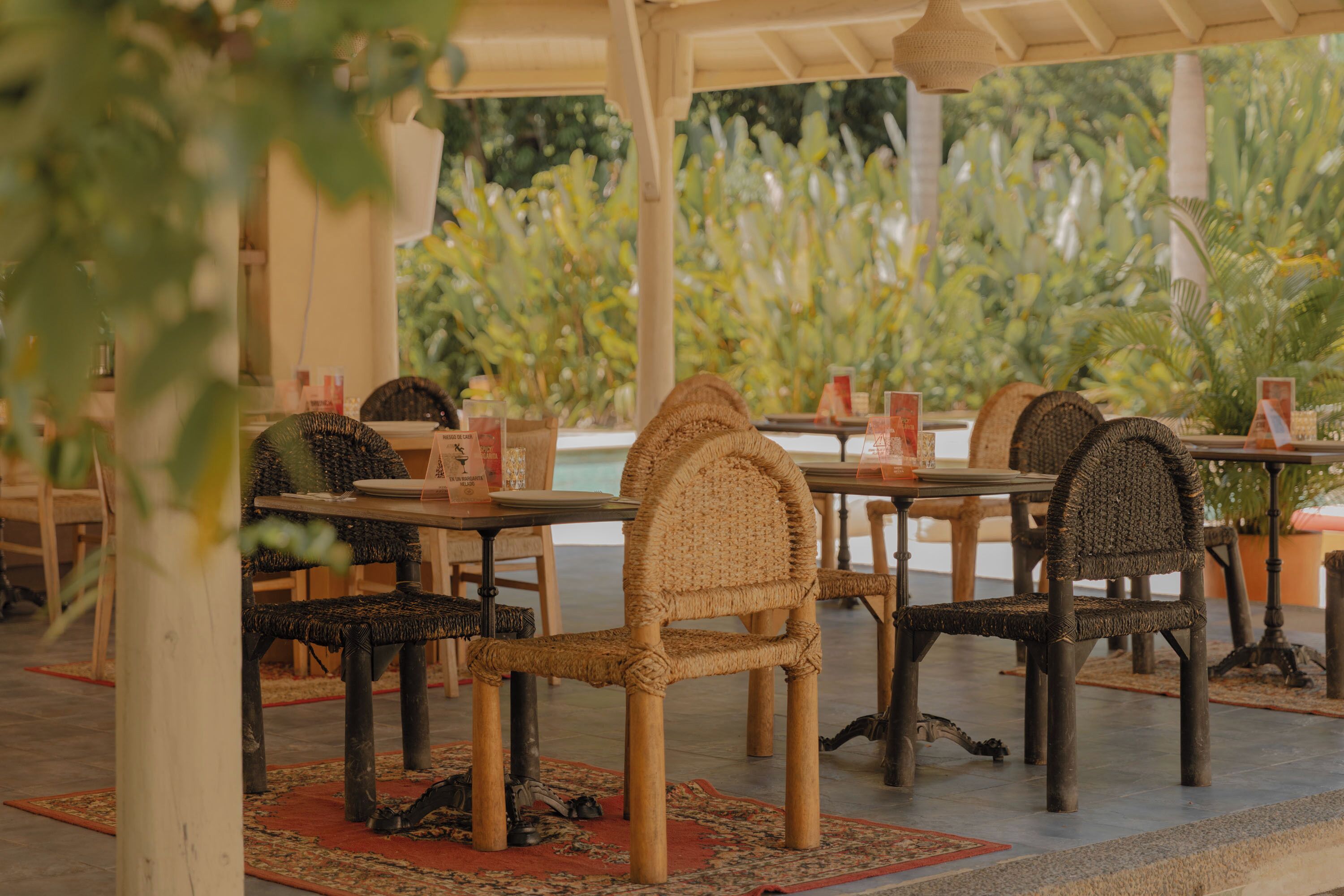 An outdoor dining area with wicker chairs and tables, a covered roof, and a view of greenery.