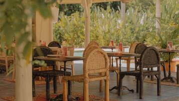 An outdoor dining area with wicker chairs and tables, a covered roof, and a view of greenery.
