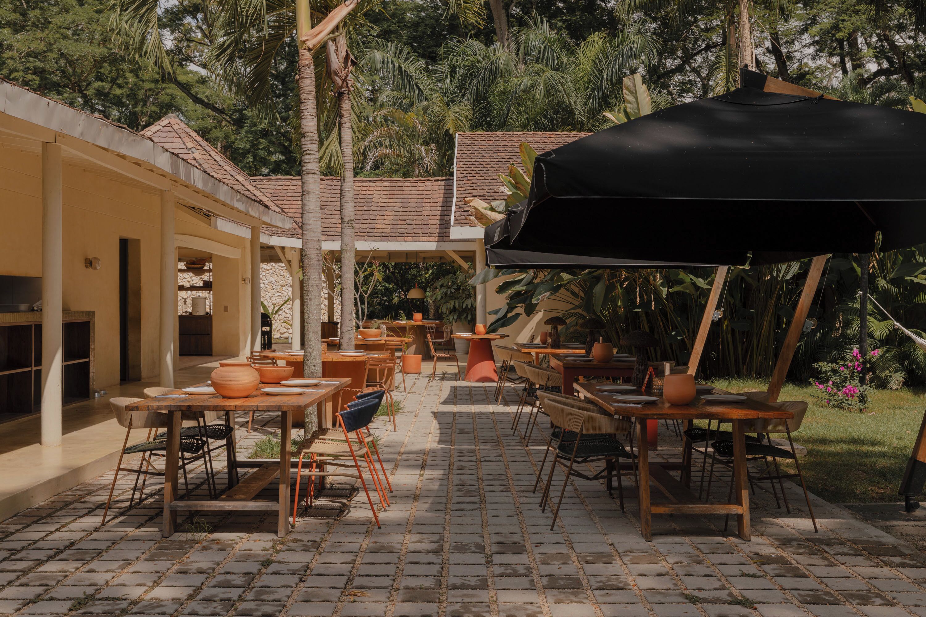 An outdoor patio with tables and chairs, surrounded by a garden and a building with a tiled roof.